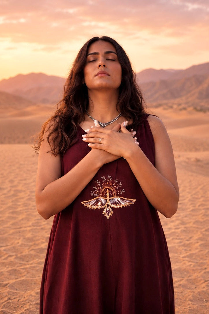 Woman in a burgundy dress standing in a desert landscape with mountains and sunset.